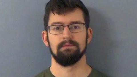 Benjamin Fields wearing a green top and glasses in a mugshot photo. He has dark hair and a thick dark beard.
