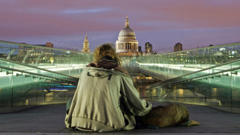 A homeless man sitting on the ground with his back to the camera, wearing a grey hoodie with a dog lying by his side. He is facing the dome of St Paul's Cathedral and on both sides of him are illuminated glass walkways. 