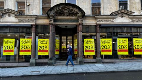 Jolly's department story with a man on the phone walking past the entrance. There are large yellow posters on every window saying 'Closing down, 20% off everything' on them.
