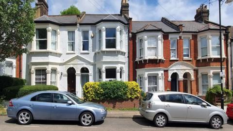 Two cars, one light blue and one silver, are parked on a residential street on a sunny day. Behind the cars is a row of semi-detached houses