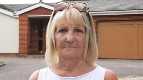 Veronica Strathie is in the middle of the image and is standing in front of a bungalow. She has shoulder length blonde hair and brown sunglasses on her head, and is wearing a white T-shirt. 