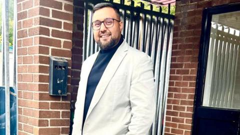 A man with short black hair, glasses, a short black beard, a white suit jacket and a dark blue top, smiles while he stands outside a door with his hands in his pockets. 