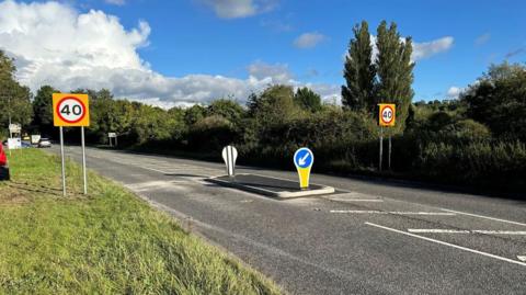 A section of the A-road where a traffic island has been newly installed in the centre of the road. On either side there are new signs signalling a 40mph speed limit. 