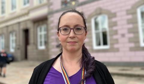 Deputy Louise Doublet in a purple top and glasses stands in front of a pink granite building, Jersey's States Assembly.