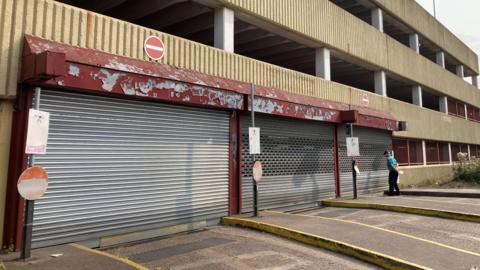 Shutters closed outside a car park, with a man looking at a sign. The building is mostly concrete.