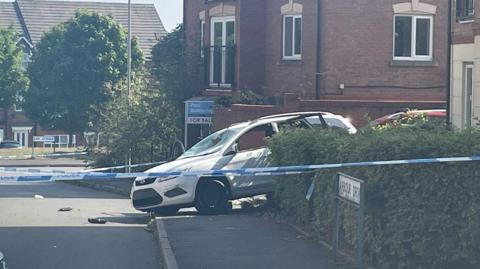 Blue and white police tape is in the foreground, near to an Arbour Drive road sign by a hedge on the right on the photo. A white car is in the centre of the image, with buildings in the background.