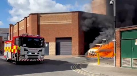 Fire engine next to van on fire. Flames can be seen coming from the van, which is parked behind the back of a commercial building