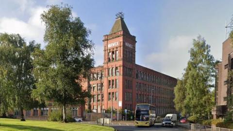 Redbrick four-storey mill with a five-storey tower that has a small mast at the top. Trees and vehicles can be seen on the roundabout in front.