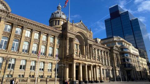 The outside of the Birmingham City Council building. It is a large brown building with pillars outside its entrance and a balcony above it. There are dozens of small windows on the front of the building  and a tower with a dome on the roof. A flag pole displays the Union Jack