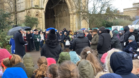 Tens of people stand gathered outside St Edmundsbury Cathedral for the event. Many wear coats and hats, while some have umbrellas, as they stand in a semi circle. 