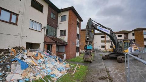 A crane is parked beside a brick apartment building. A pile of debris is on the ground to the left. 