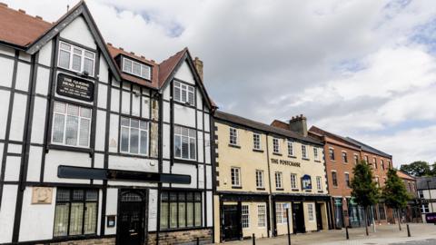 The Queens Head, Postchaise and Monaco buildings are three-storey buildings that stand side-by-side. The Queens Head has a black and white timber design on the exterior. The Postchaise is painted beige and the Monaco is a red-brick building with black doors.