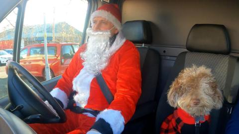 Bowie the labradoodle sits on the front passenger seat of his master's van. Bowie is dressed in a festive red-and-black dog jacket. Meanwhile the dog's owner, Ollie Foster, is pictured at the wheel, wearing a full red-and-white Santa suit (including long white beard)