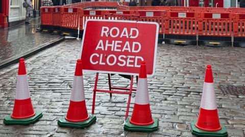 A red and white road sign which reads: "Road ahead closed.". It is placed on a cobbled road with four red and white cones set out in front of it. Behind it, there is a line of orange safety barriers along the width of the road also.
