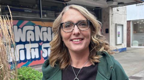 Lindsey Atkins standing in Kettering high street. She is in a green hooded jumper and wears eye glasses with large frames. Her hair is shoulder length and blonde and she is smiling at the camera.