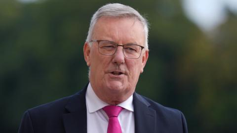 Nesbitt is wearing black thin framed glasses, a black suit and a pink tie. He has short greyish hair and the background is blurred.