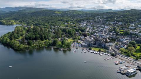 An aerial view of Bowness-on-Windermere, a lake shore town in the Lake District National Park. There are lots of boats moored in the marina and the town stretches out beyond it, surrounded by trees. In the distance are hills and mountains.