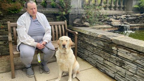 Lee sitting on an outdoor bench outside a hotel next to pond with his golden retriever guide dog on a lead.