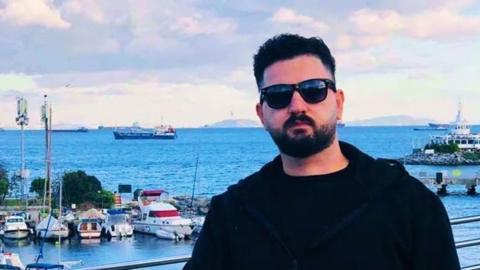 Kardo Jaf pictured in a coastal setting. He has short black hair and facial hair, and wears black sunglasses and a black top. Behind him is a large body of water where several boats are docked in a marina, and 