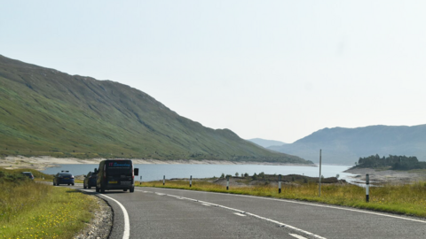 A grey road with white road markings can be seen in the foreground with grass either side. Four vehicles, including a black van, are on the road. In the background, a body of water is present between two large black and green hills, with a clear light blue sky.