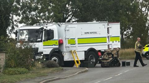 White truck with yellow and blue stripe. Bomb disposal written on it. There is a yellow ladder, a bomb disposal robot, a police car and a man in police bomb disposal officer. There are trees, bushes, a road and a path.
