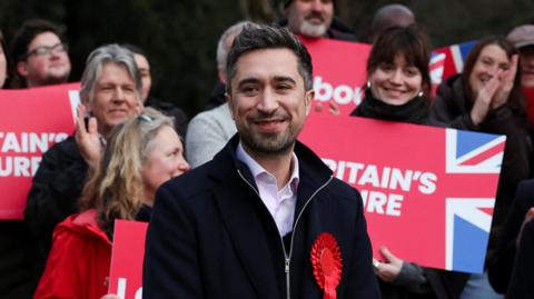 Damien Egan wearing a red rosette, stands in front of a group of people, some are holding placards reading: "Britain's Future"