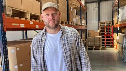 A man wearing an open chequered shirt, and a white t-shirt, is standing inside a warehouse.