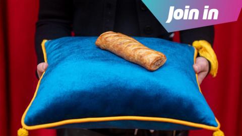 A waxwork of a Greggs sausage roll sits on a blue cushion
