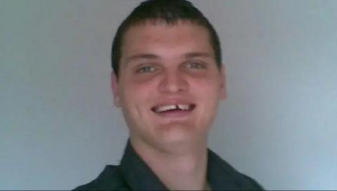 Kyle Marshall has blue eyes and short cropped dark hair. He is wearing a black shirt and is smiling at the camera. Behind him is a plain grey background.