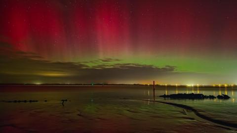 Mud flats at Great Yarmouth in shadow with yellow, green and pink skies of the Northern Lights. Lights on the edge of the shoreline are in the distance.
