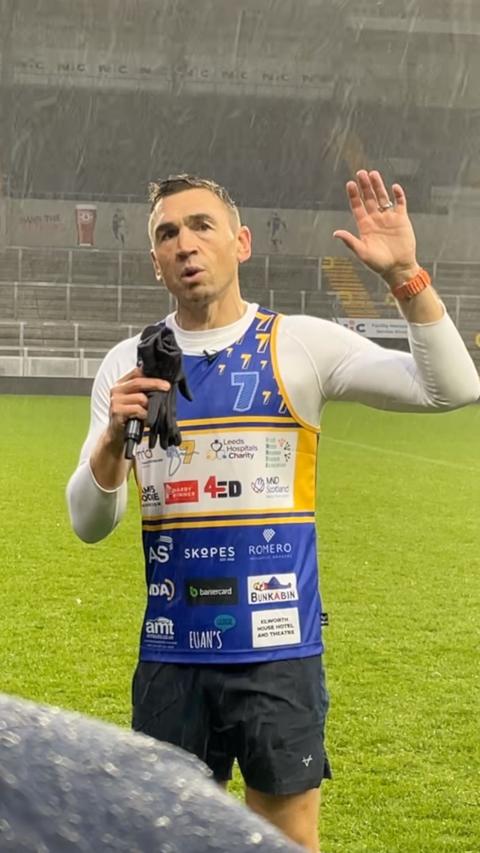 Kevin Sinfield with microphone and gloves in one hand raises his other hand during a speech at Headingley Stadium.