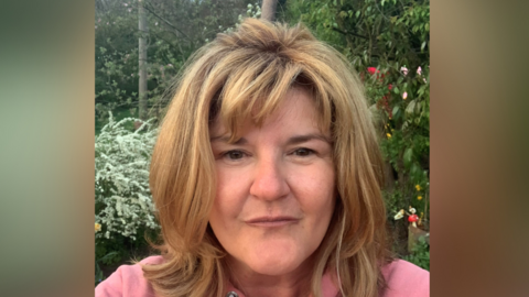 A selfie image of Nichola Greenfield. She has shoulder length hair and is wearing a pink hoody. She is standing in front of some trees. 