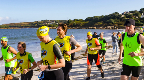 The picture shows a group of people taking part in a swim-run race. The participants are dressed in wetsuits and brightly coloured vests with numbers, and some are wearing swim caps and goggles. In the background, you can see a scenic coastal landscape with hills, houses, and the ocean.