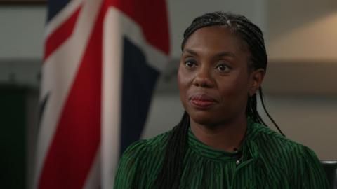 Kemi Badenoch sits in front of a British flag. She has long, braided hair. She is wearing a brown and black striped top.