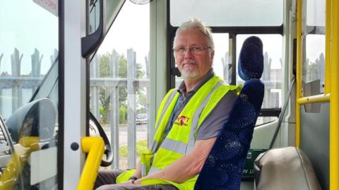 A man with a beard and grey hair seated behind the steering wheel of a minibus. He is wearing a yellow hi-vis vest. The vehicle is equipped with bright yellow grab rails.