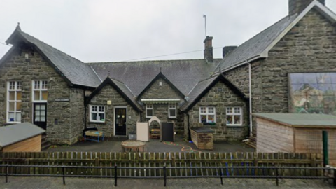 A general view of the primary school, a grey stone building with a pitched slate roof. It has white window sills and black doors, with a yard in front of the school and a fence in front of it.