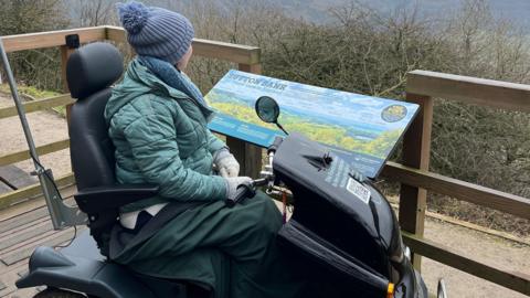 A young woman with a woolly hat sits on a large black off-road mobility scooter. She wears a green puffed jacket and a blue and white striped jumper. She is parked on a raised wooden viewing platform overlooking a countryside scene.