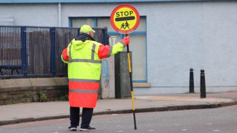 A lollipop man is wearing bright clothing and holding a crossing patrol pole in a Scottish street.