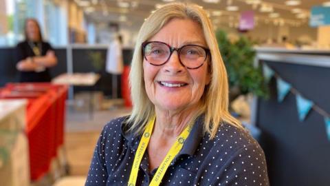 A woman with long blonde hair, glasses, a yellow lanyard and a blue top with white spots, smiles as she stands in a store. There are people, bunting and lights all blurred in the background. 