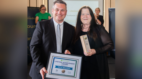 Professor Julia Williams receiving her Lifetime Achievement Award from College of Paramedics president Jon Price. Jon is holding the certificate and is wearing a black suit and silver tie. Julia, who has long black hair, is wearing a black dress. 