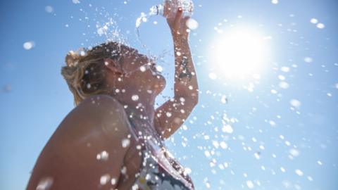 Woman pours a bottle of water over hear head as she stands in sun rays.