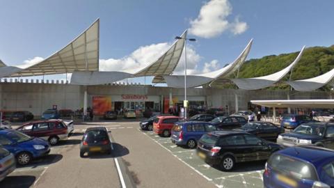 A photo of the exterior of Sainsbury's at Marsh Mill in Plymouth that is taken from the car park and there are cars parked in the foreground.