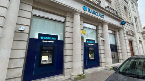The exterior of a bank. Two ATMs are visible. On the left of the image, the sign above the ATM says 'Barclays Jersey Currency. On the right of the image, the sign above the ATM says 'Barclays English Currency'. Above the ATMs, a bigger sign, in light blue, says 'Barclays'.