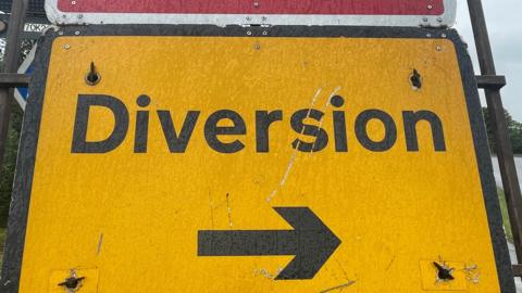 A bright yellow road sign which says "Diversion" in black with a black arrow underneath. It is resting on an verge, with a road on the right. In the far distance beyond the sign is a lorry.