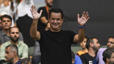 A man with short dark hair and wearing a black T-shirt stands up from a crowd of people, waving with both hands in the air.