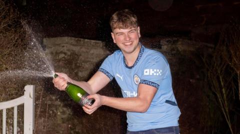 James Clarkson in a garden shaking a bottle of Champagne which is spraying out of the bottle