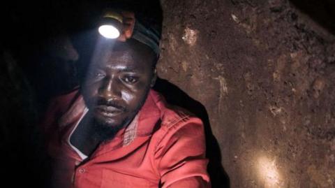 A man in a red overall in an underground mine looks downward as a torch on his helmet illuminates what's in front of him.