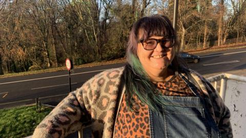 A woman has long black hair with a green streak. She is wearing a patterned cardigan, a leopard print top and dungaree dress and is standing on a council flat veranda with woodland in the background.