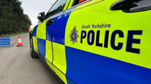 A South Yorkshire Police car with bright yellow‑and‑blue markings parked on a road, with a traffic cone and a blue “Police Accident” sign set up nearby, against a backdrop of trees.