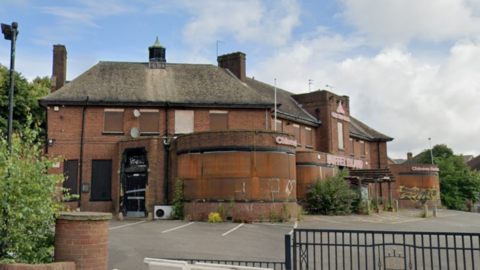 A large red brick building can be seen which is boarded up. It is quite run down and has a large car park in front of it with a fence on one side.